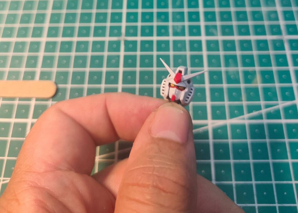 A close-up of a hand holding a tiny Gundam model headpiece, painted in white, red, and grey with yellow eyes and V-fin antennae. The background shows a green cutting mat with a grid pattern, commonly used for scale modelling.