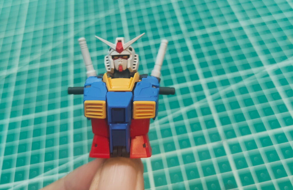 A close-up of a hand holding the partially assembled torso and head of a Gundam model kit. The figure has a white head with red and grey details, blue chest armour with yellow vents, red lower torso, and two grey cannons on its back. The background is a green cutting mat with a grid pattern.