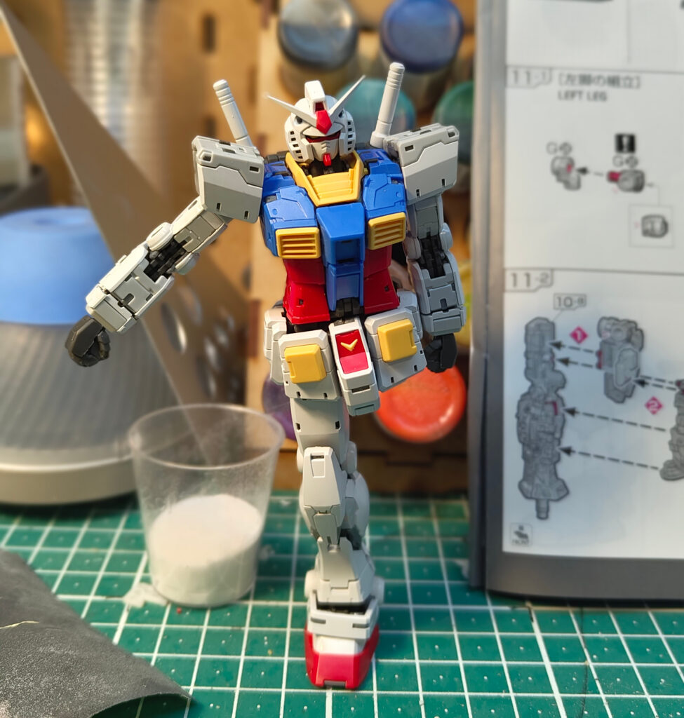 A partially assembled RX-78-2 Gundam model kit standing on one leg in a dynamic pose on a green cutting mat. The model has its torso, arms, and one leg attached, painted in its classic colours of white, blue, red, and yellow. In the background are hobby supplies, paints, and the model’s instruction manual.