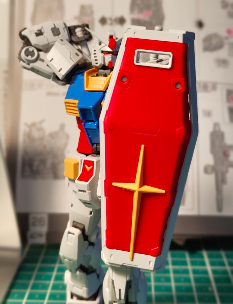 A close-up of an RX-78-2 Gundam model kit holding its large red shield with a yellow cross emblem. The Gundam is posed with one arm raised behind its head, and the shield covers most of its body. The background shows an instruction manual slightly out of focus on a hobby workbench.