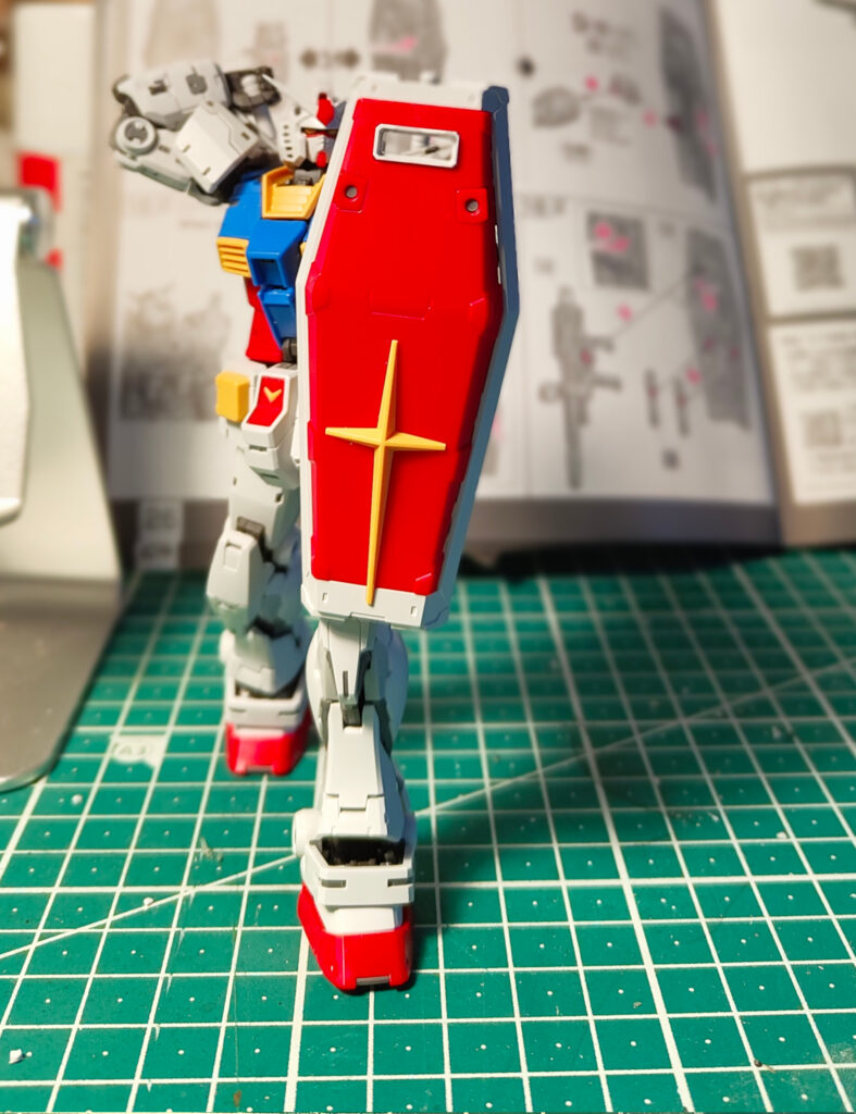 A completed RX-78-2 Gundam model kit standing on a green cutting mat, holding its large red shield with a yellow cross emblem. The model’s white, blue, red, and yellow armour is visible, and the background shows an open instruction manual and hobby workspace.