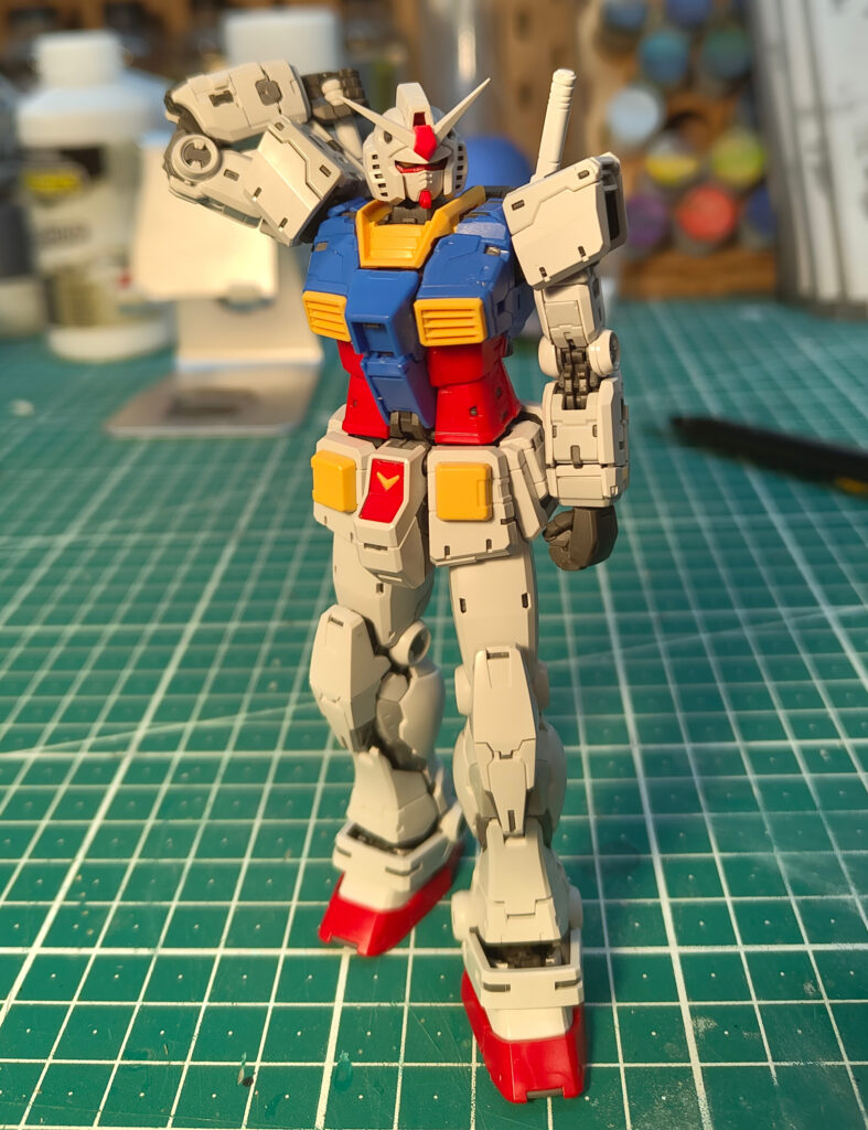 A completed RX-78-2 Gundam model kit standing on a green cutting mat, posed with one arm raised behind its head in a casual stance. The model features its classic red, blue, yellow, and white colour scheme with red feet. The background shows hobby tools, paints, and supplies on a workbench.